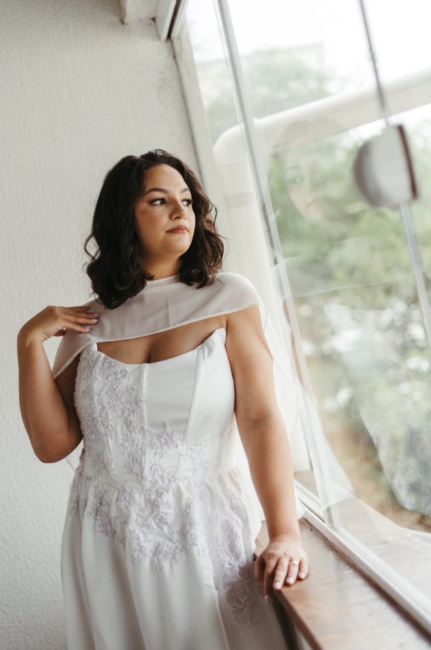 Modelo com vestido de noiva branco e capa de tule, junto à janela